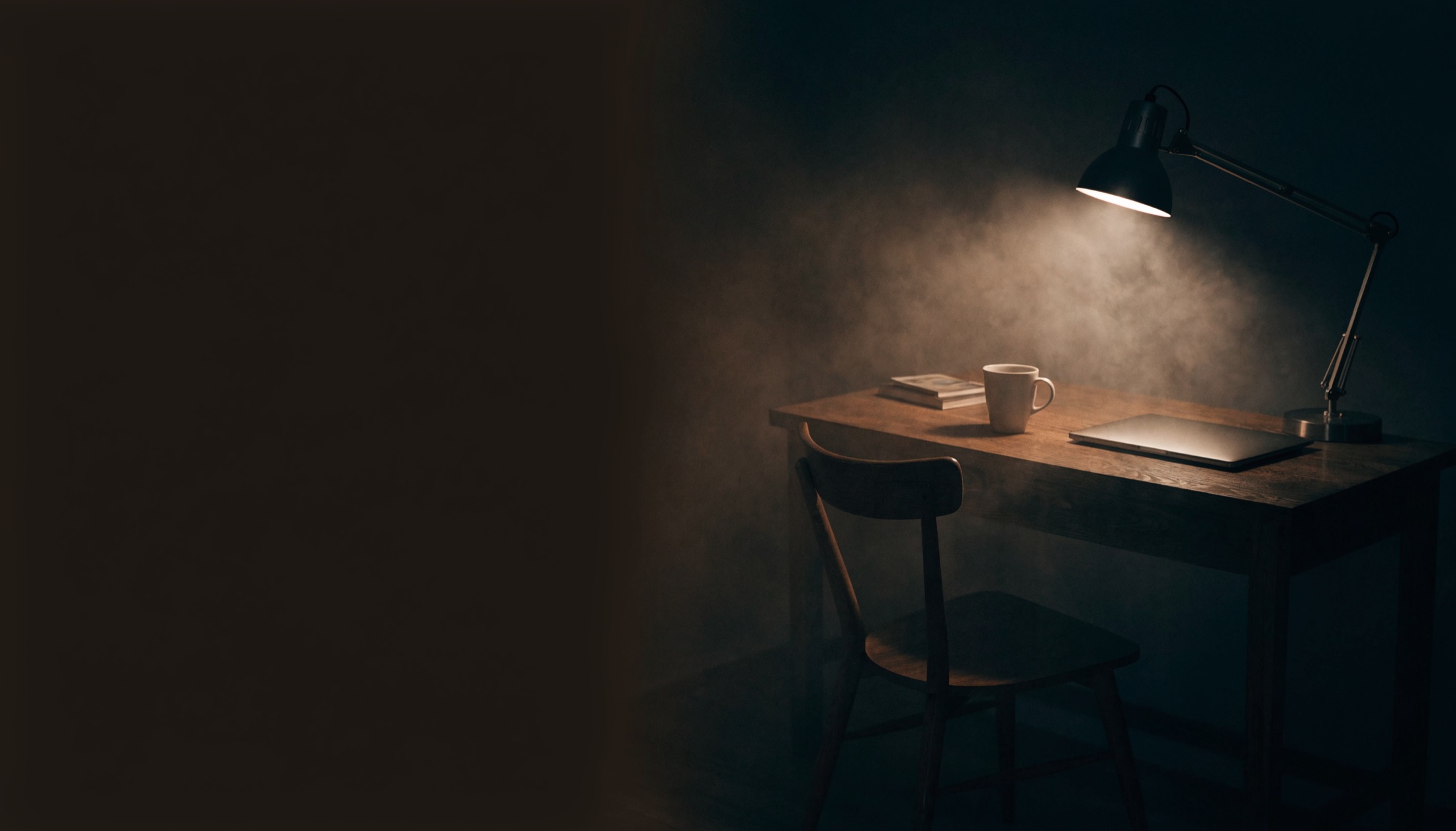A single desk lamp still on in a dark room at night, lighting an empty chair and a laptop